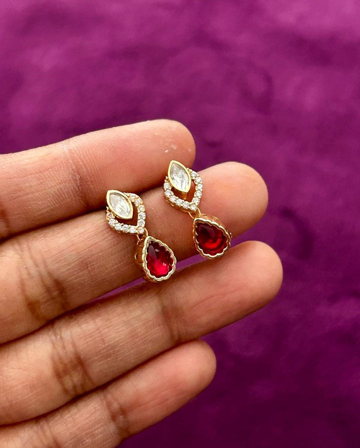 Gold earrings with red gemstones held between fingers against a purple background