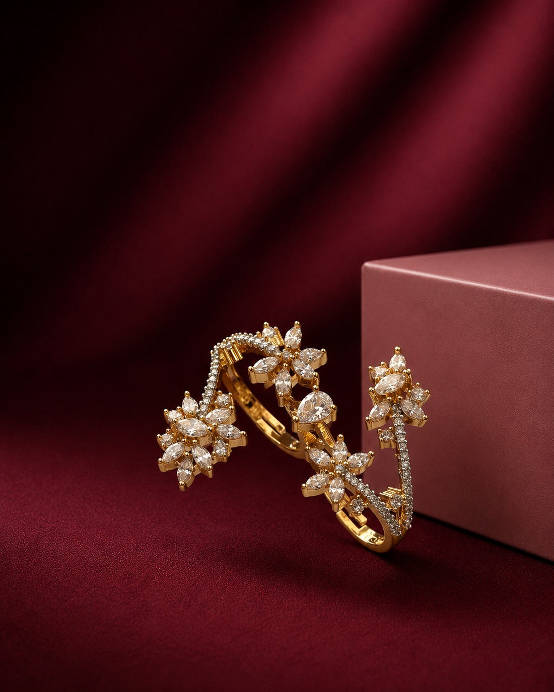 Gold floral cuff bracelet with pavé-set clear stones, displayed on a dark red satin fabric next to a dusty rose jewelry box.