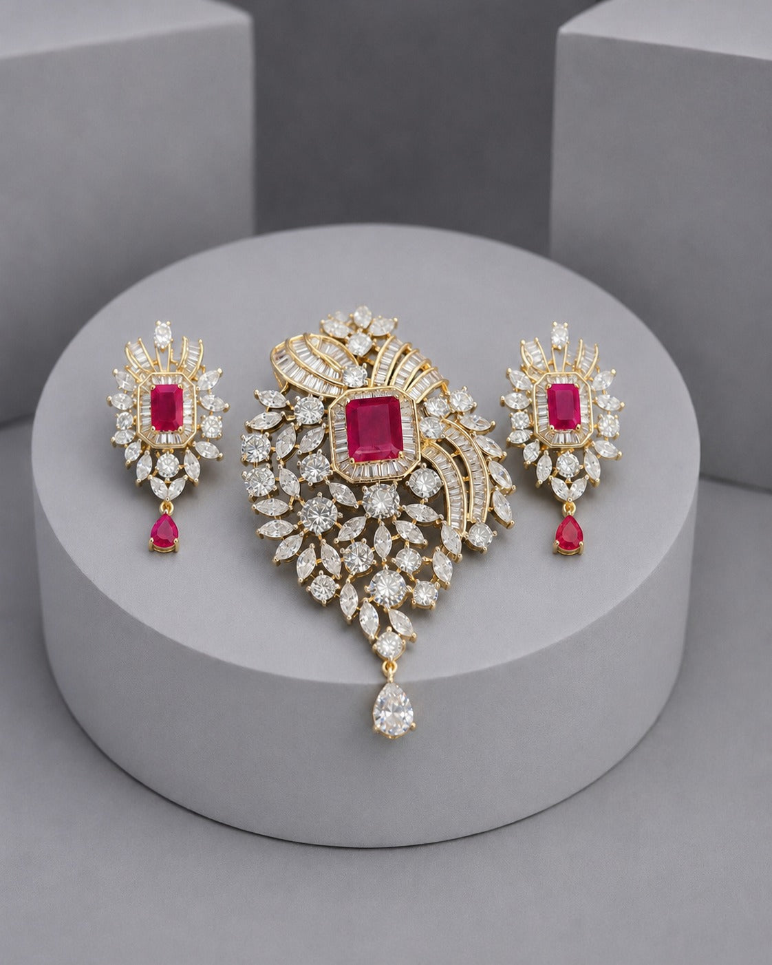 Gold jewelry set featuring a large central brooch and two matching drop earrings, decorated with clear crystals and square pink gemstones, displayed on a round gray pedestal.