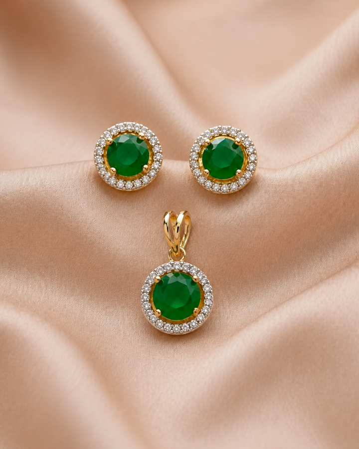 Gold jewelry set featuring a pendant and matching stud earrings with round green gemstones surrounded by clear crystal halo settings, displayed on a draped beige satin fabric.