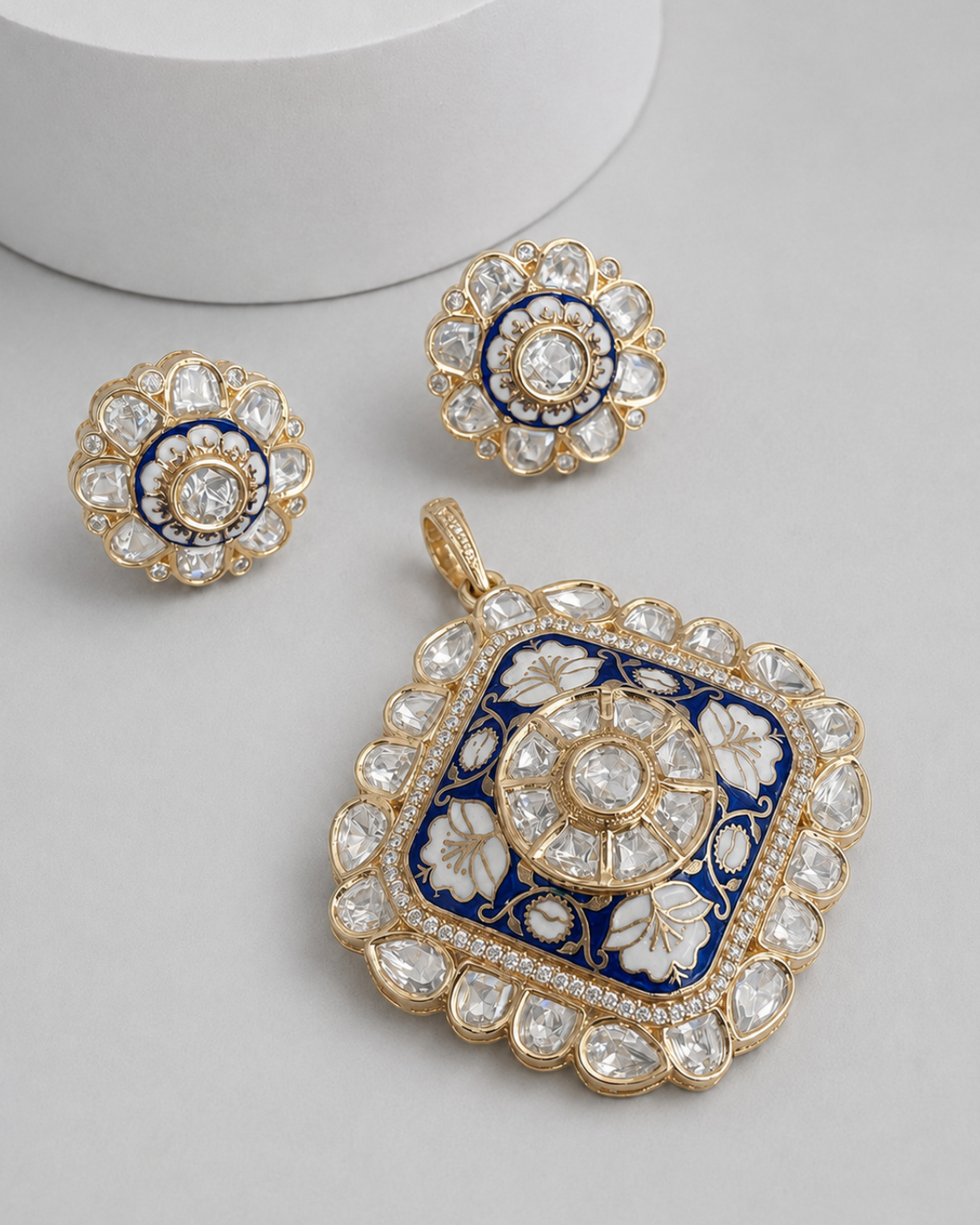 Gold jewelry set featuring a square pendant and two matching round earrings, decorated with white floral enamel, blue accents, and clear stones on a light gray background.
