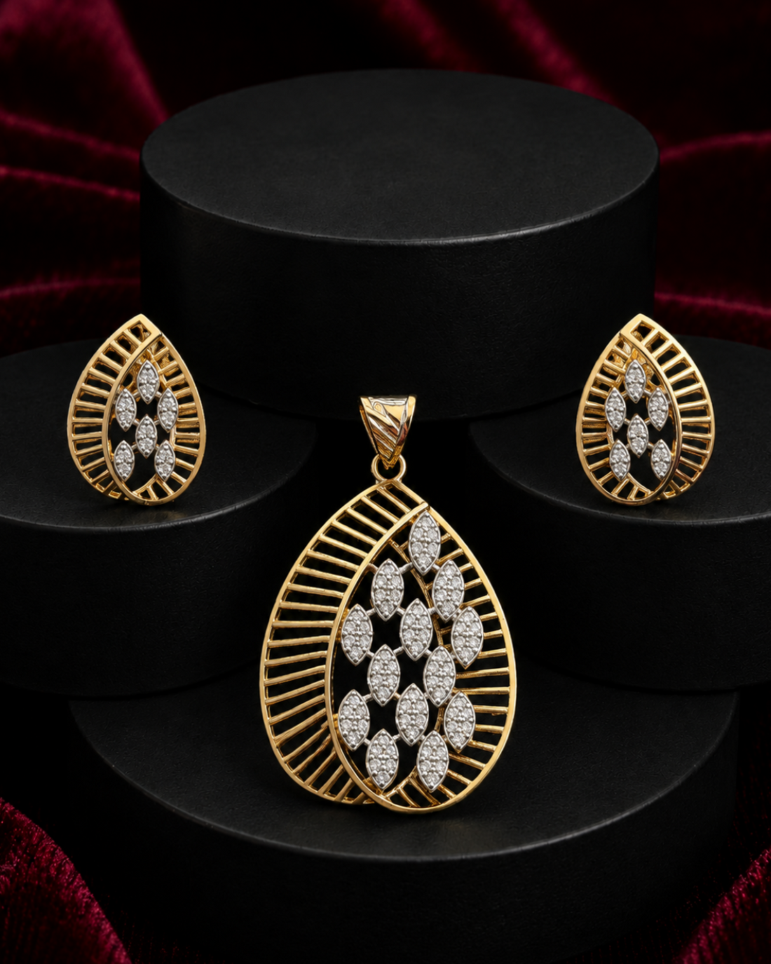 Gold jewelry set featuring a teardrop-shaped pendant, matching earrings, and a small box, all adorned with sparkling clear stones in a leaf-like pattern, displayed on black cylindrical pedestals against a dark red velvet background.