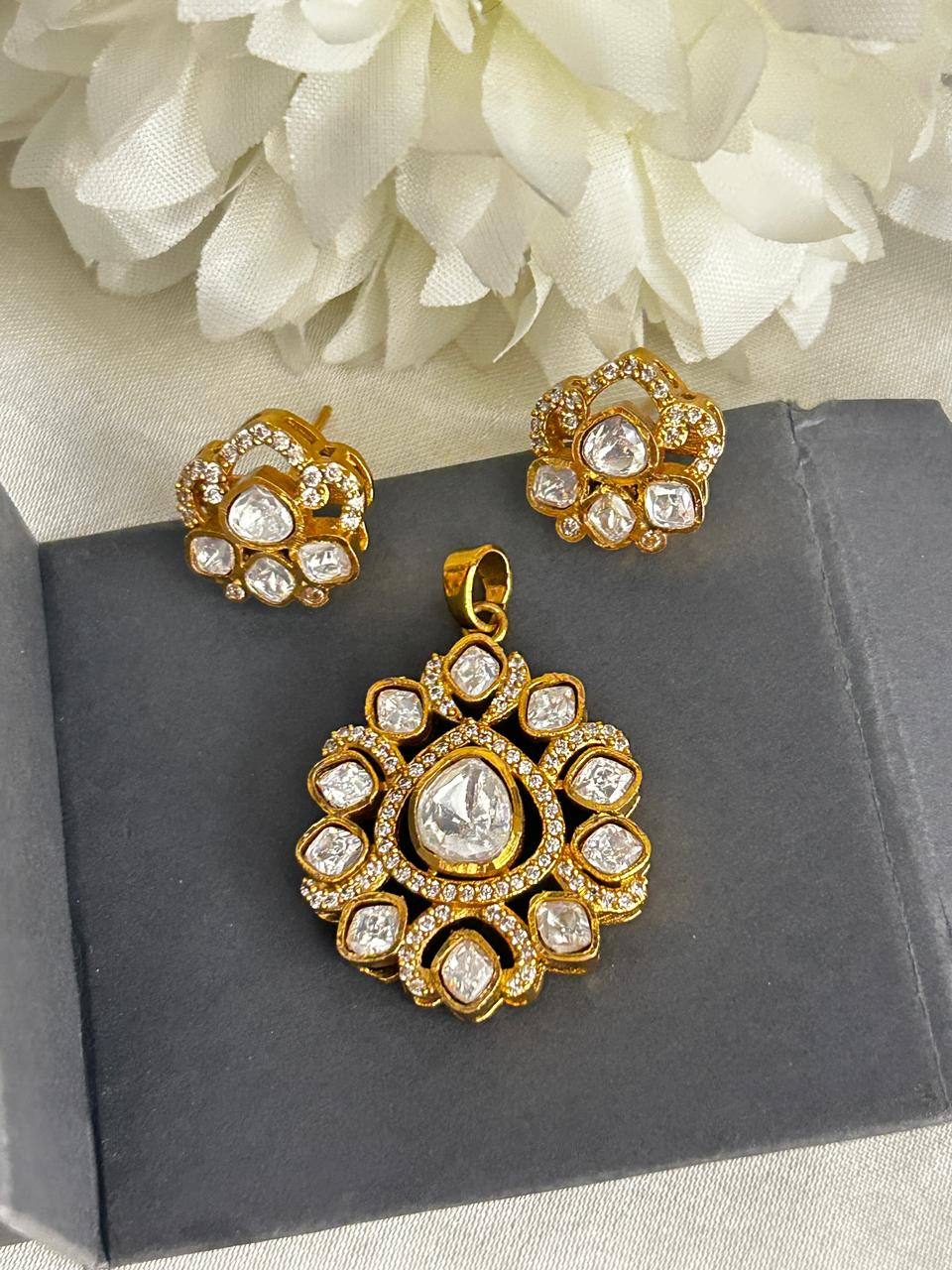 Gold jewelry set with pendant and earrings on a dark surface with white flowers in the background