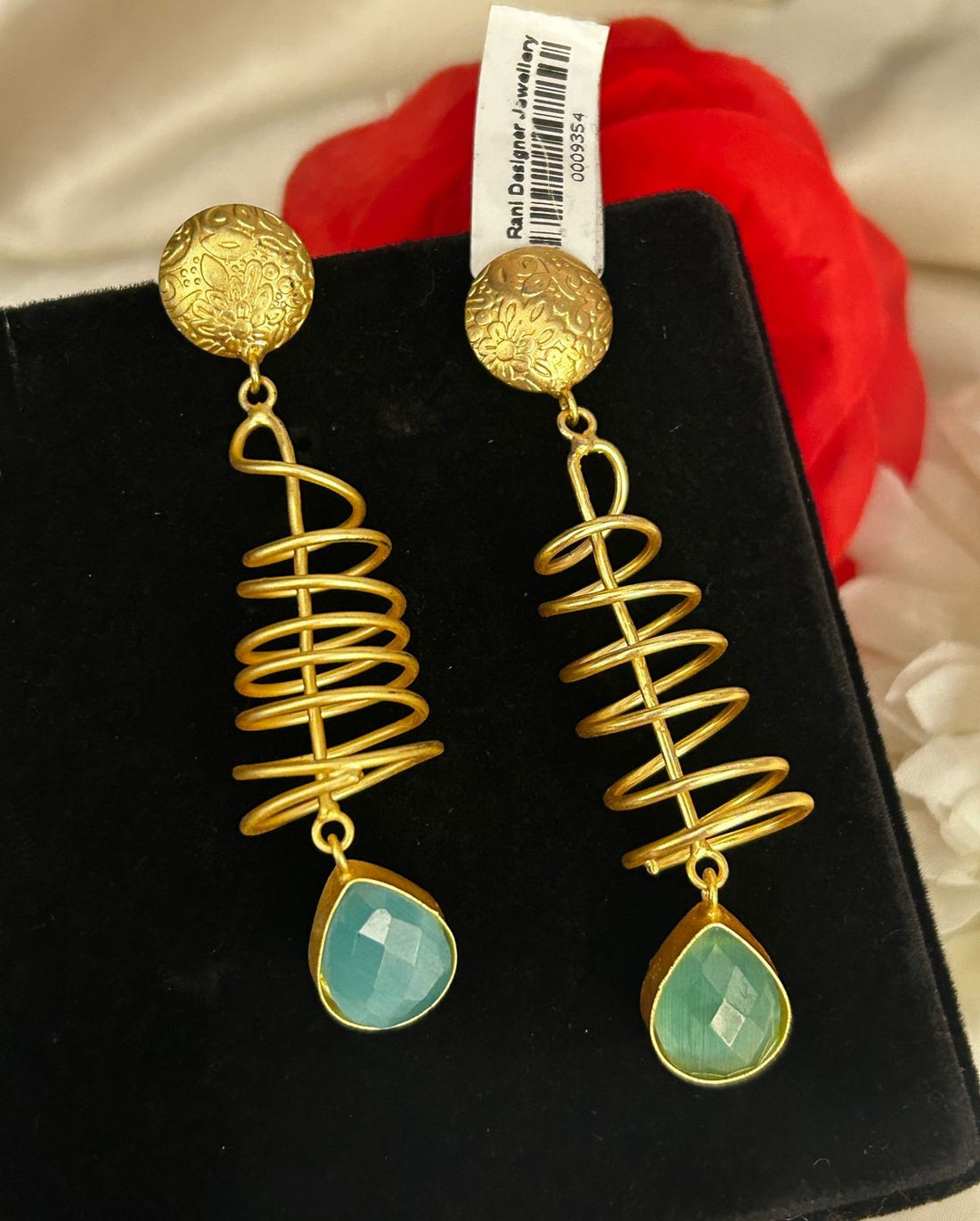 Gold spiral earrings with green teardrop charms on a black stand with a red flower in the background.
