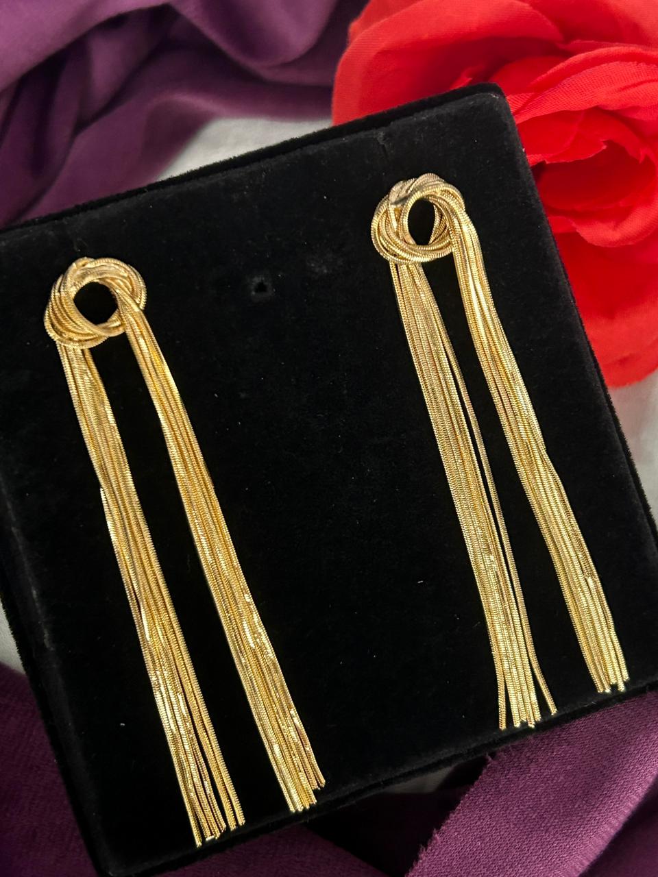 Gold tassel earrings on a black display box with a red flower in the background.