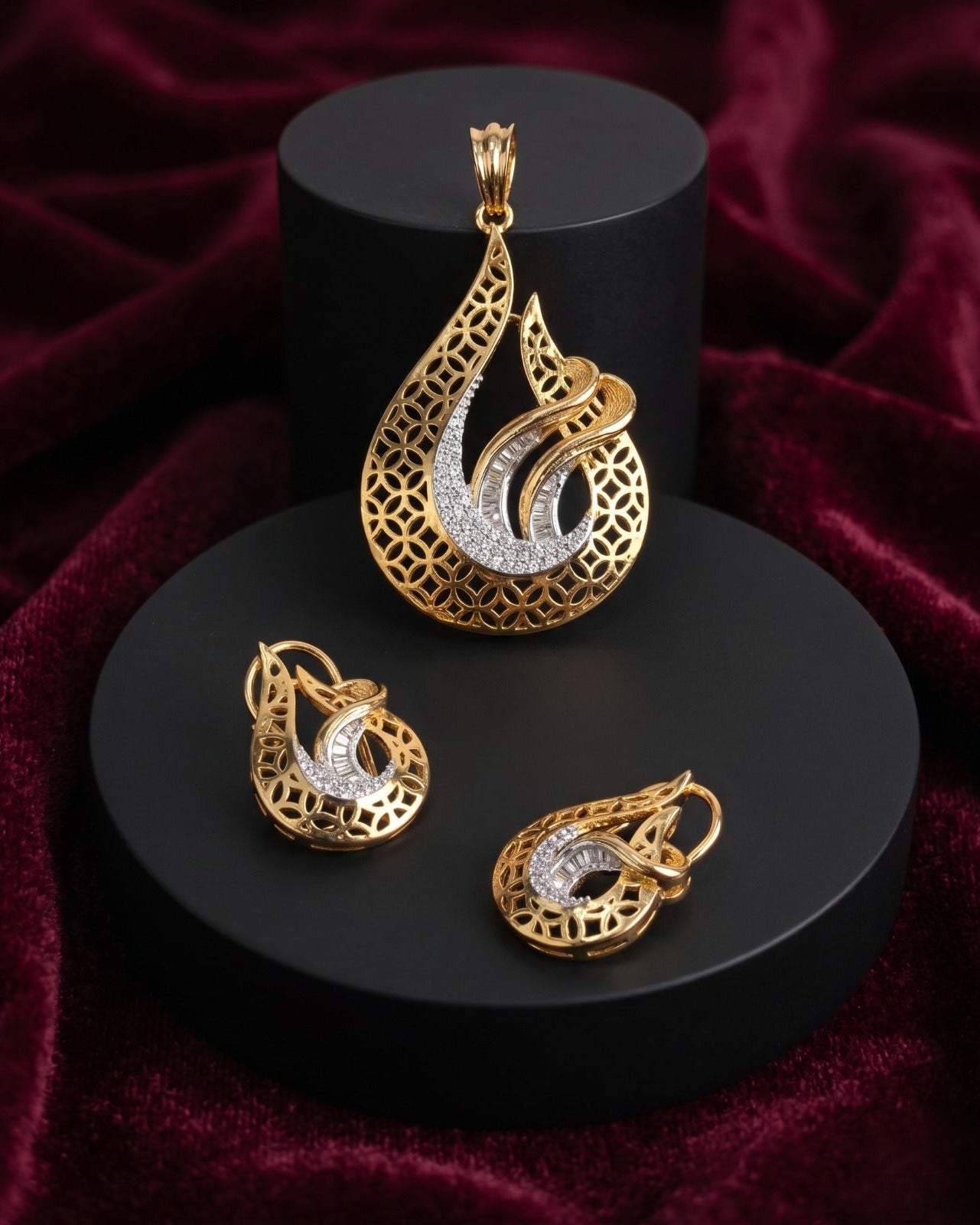 Gold teardrop pendant and matching earrings with intricate filigree detailing and a curved silver inlay, displayed on black cylindrical pedestals against a deep burgundy velvet background.