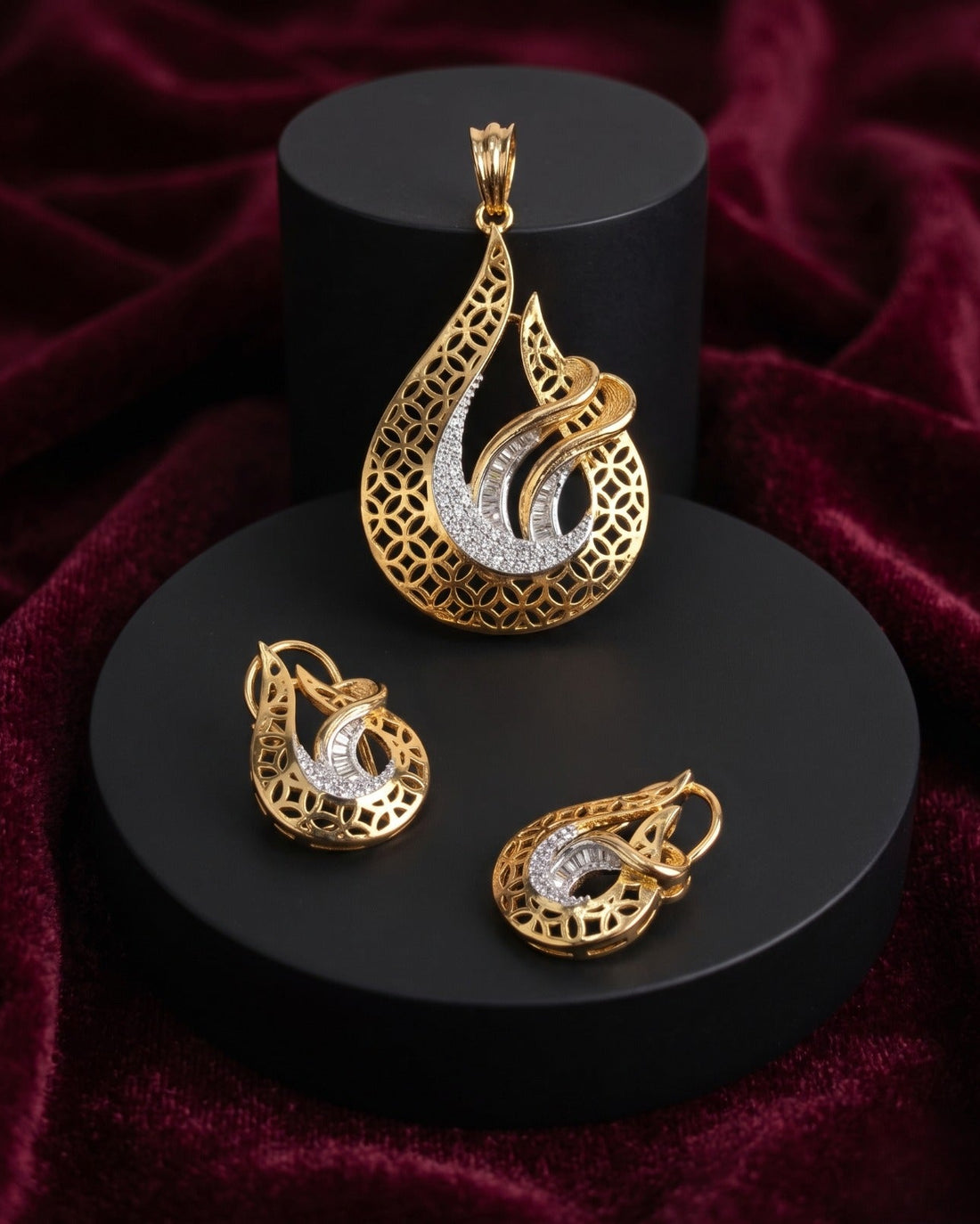 Gold teardrop pendant and matching earrings with intricate filigree detailing and a curved silver inlay, displayed on black cylindrical pedestals against a deep burgundy velvet background.