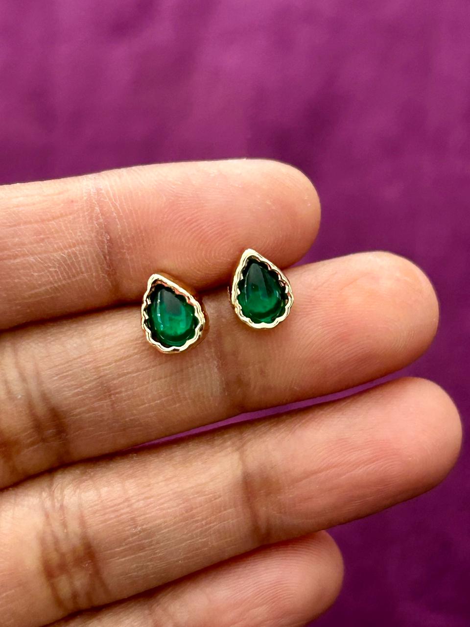 Gold earrings with green gemstones held between fingers against a purple background