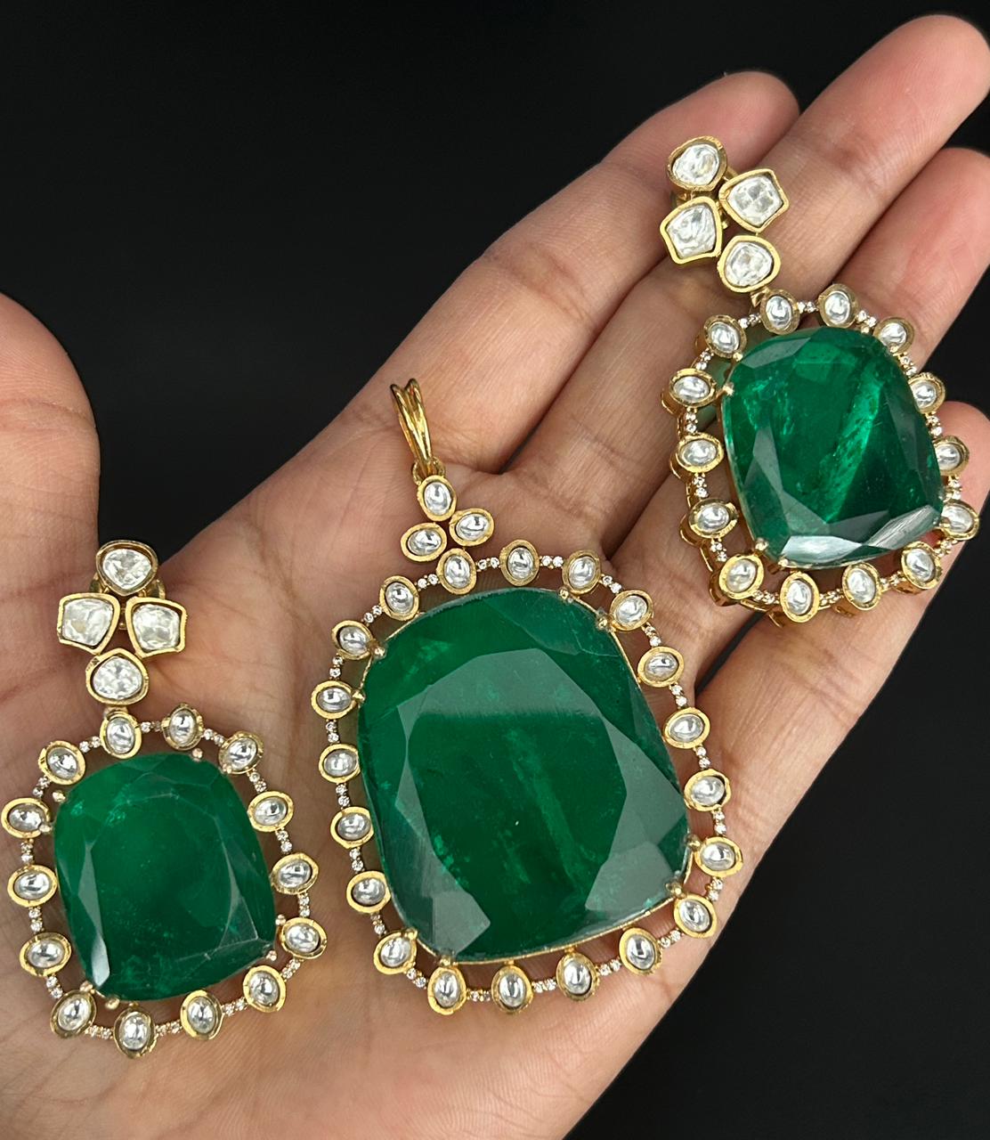 Green gemstone earrings with gold and pearl accents held in a hand against a black background