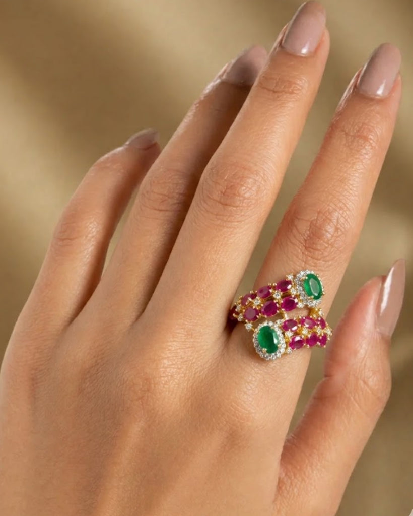 Hand wearing a ring with red and green gemstones against a beige background