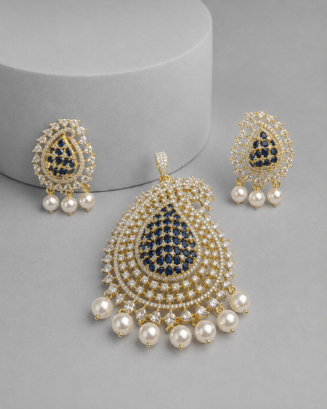 Gold-plated traditional Indian jhumka earrings and a matching pendant necklace set, featuring a central navy blue teardrop inlaid with small clear stones, surrounded by rows of white faux pearls and dangling white pearls, displayed on a gray background.