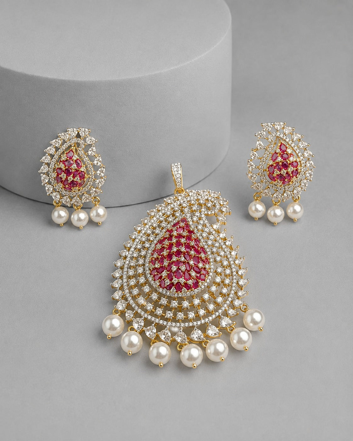 Gold-toned traditional Indian jewelry set featuring a teardrop pendant and matching earrings, embellished with white faux pearls and red gemstone centers, displayed on a gray background.