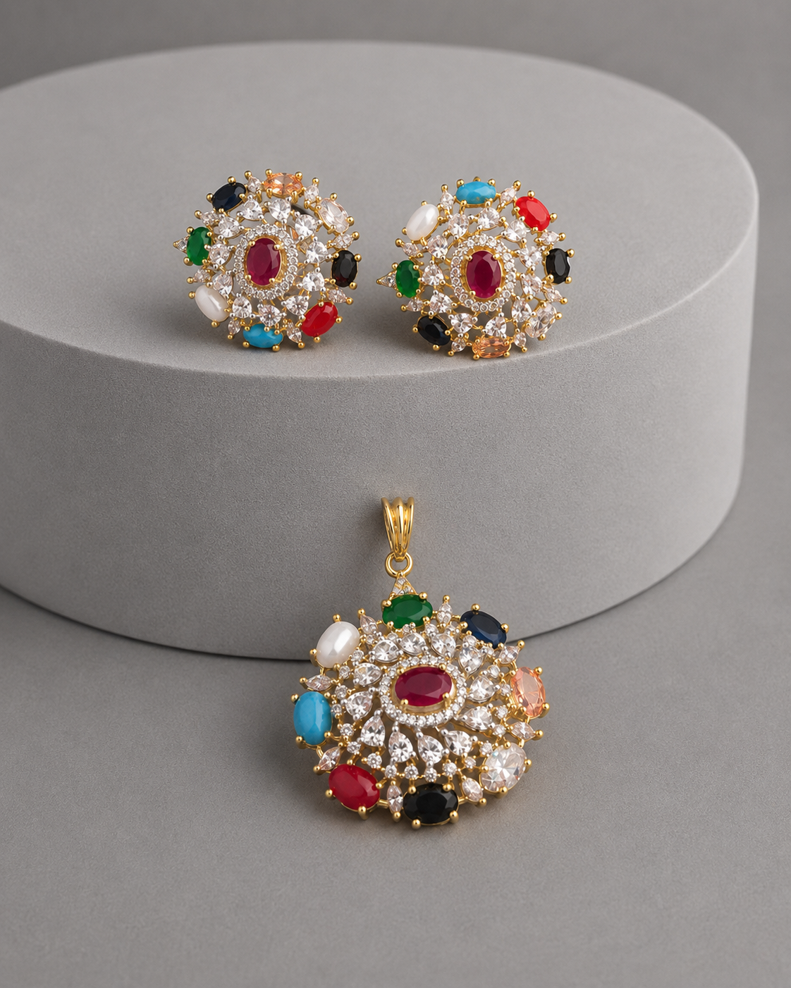A gold-tone jewelry set featuring a matching pendant and stud earring, both adorned with a colorful array of faux gemstones including red, blue, green, black, and white, displayed on a gray cylindrical pedestal.