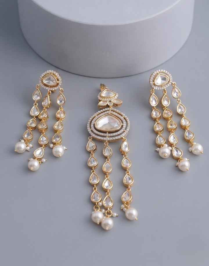 Gold-toned traditional Indian chandelier earrings with white faux pearls and clear stones, displayed on a gray surface with a white cylindrical prop.