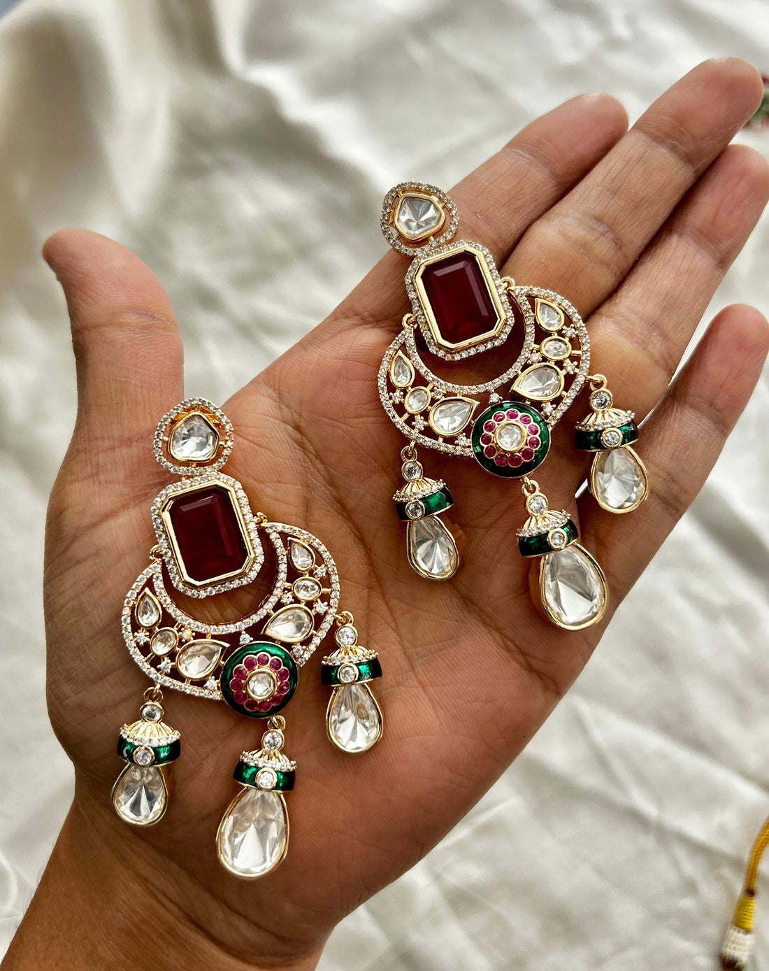 Ornate earrings with red and green stones held in a hand against a neutral background