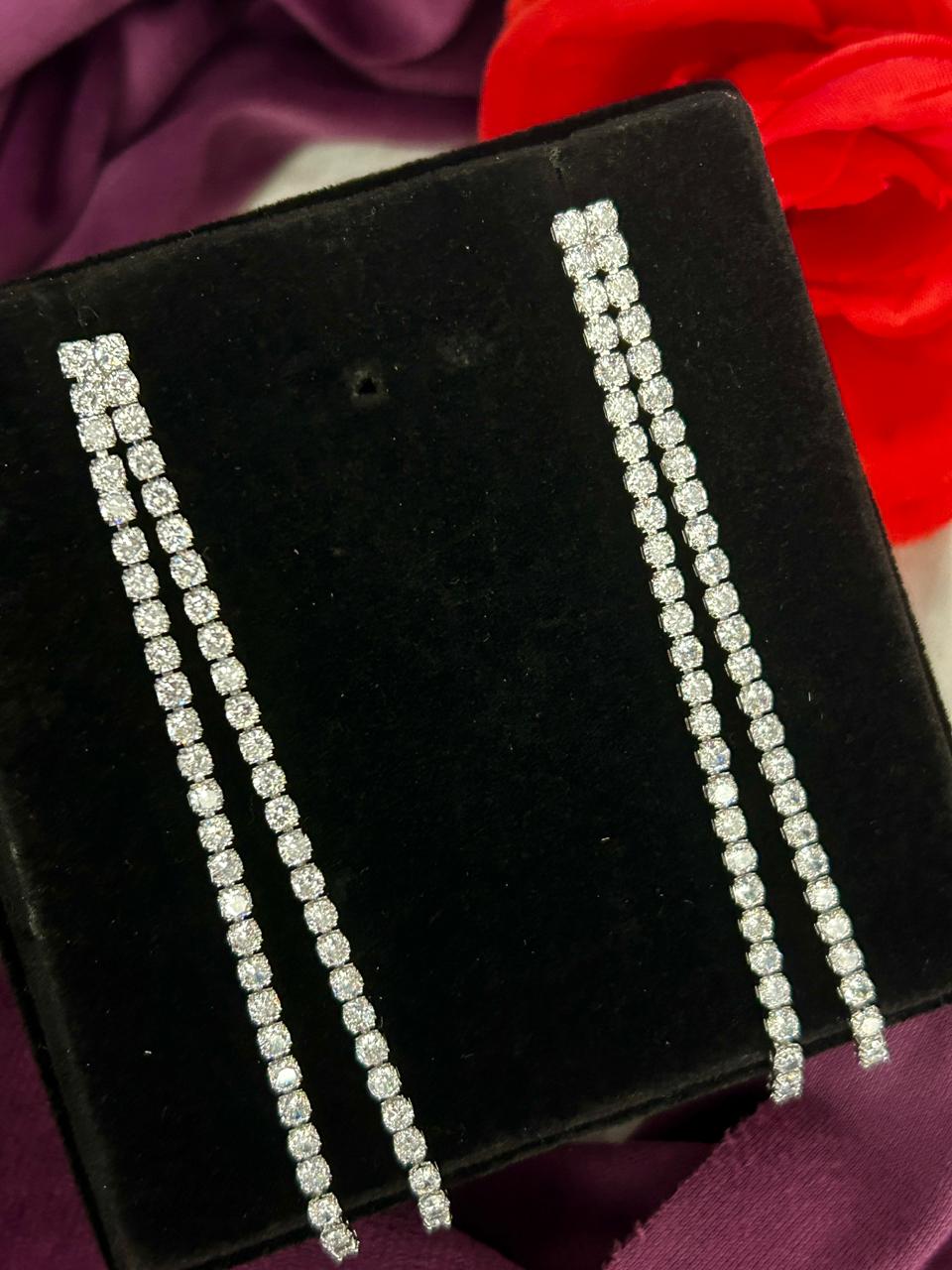 Pair of diamond earrings on a black velvet stand with a red flower in the background.