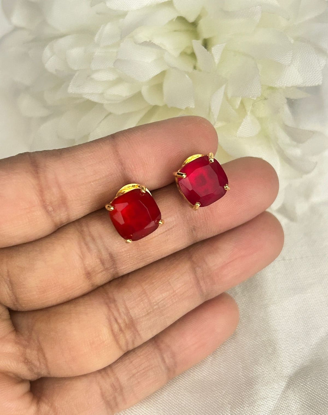 Red earrings with gold accents held between fingers against a white floral background