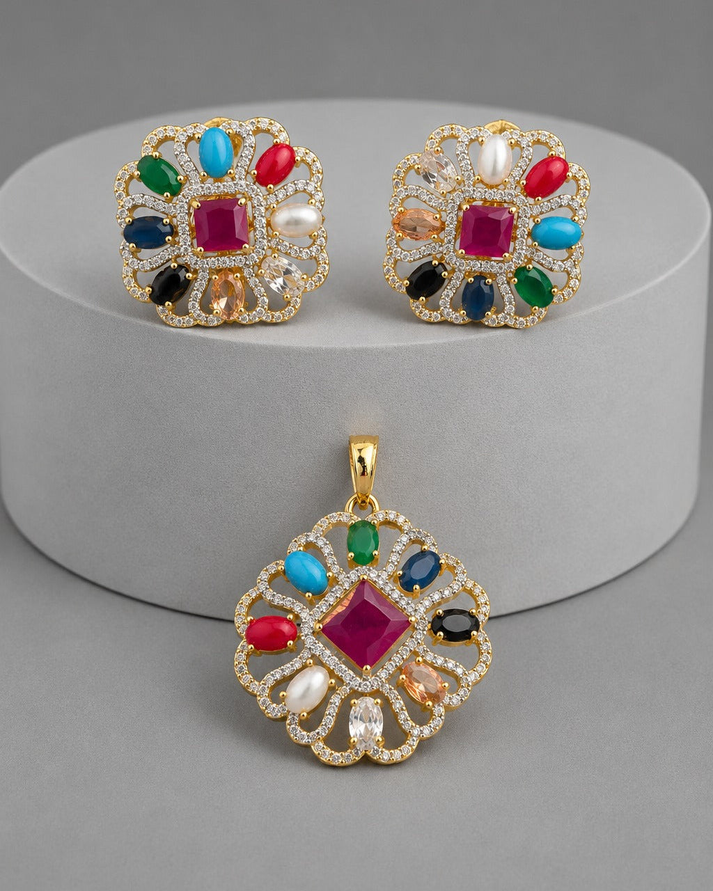 Gold jewelry set featuring a matching pendant and earrings with a colorful gemstone pattern, displayed on a gray cylindrical pedestal.