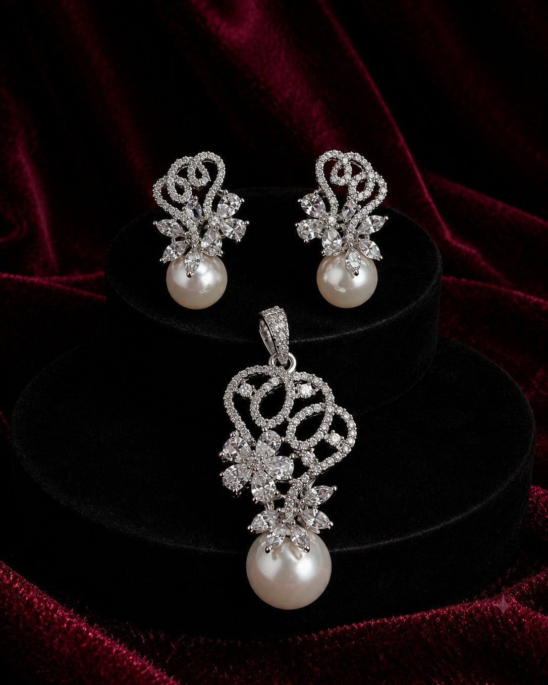 Silver-toned pearl drop earrings and matching pendant set with pavé crystal filigree, displayed on black velvet stands against a deep burgundy velvet background.