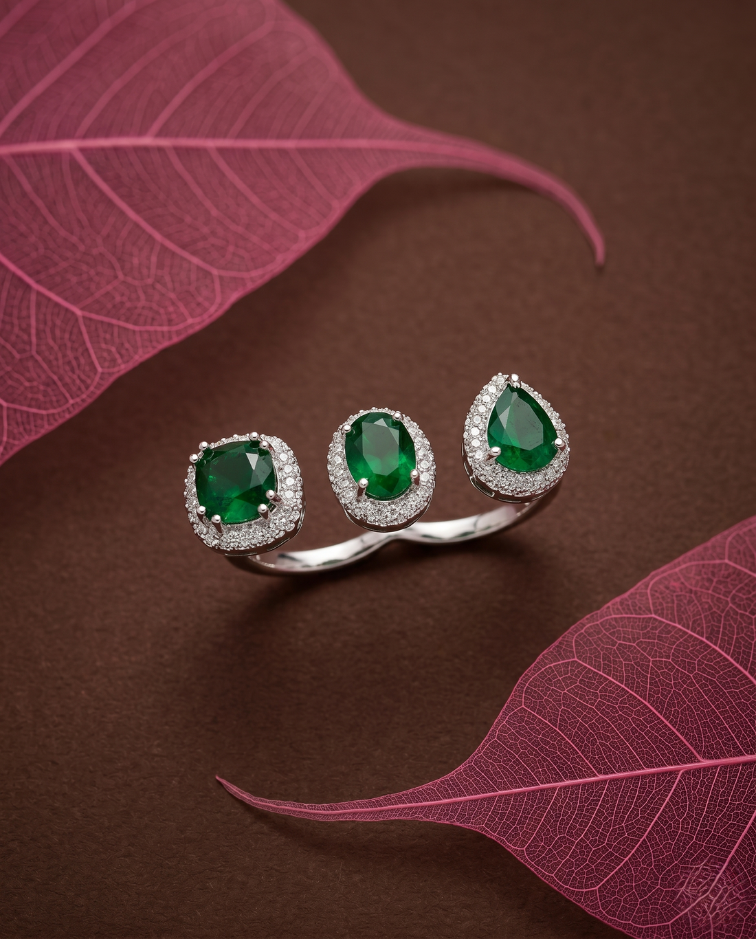 Silver ring with green gemstones on a brown surface with pink leaves 

