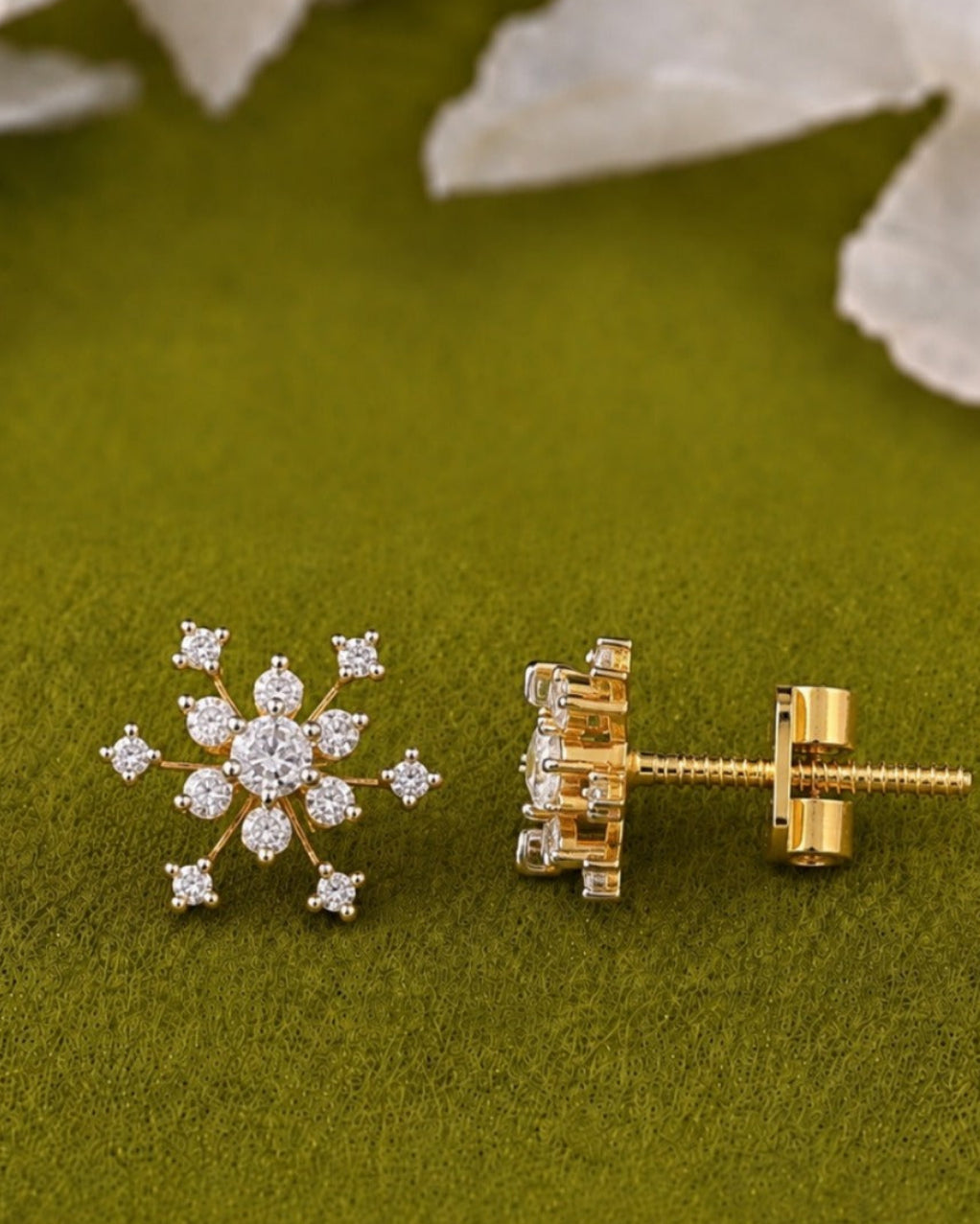 A pair of gold stud earrings, one shown from the front as a snowflake-like floral cluster with clear stones and the other from the side showing the post and butterfly back, displayed on a textured olive green surface with blurred white flowers in the background.