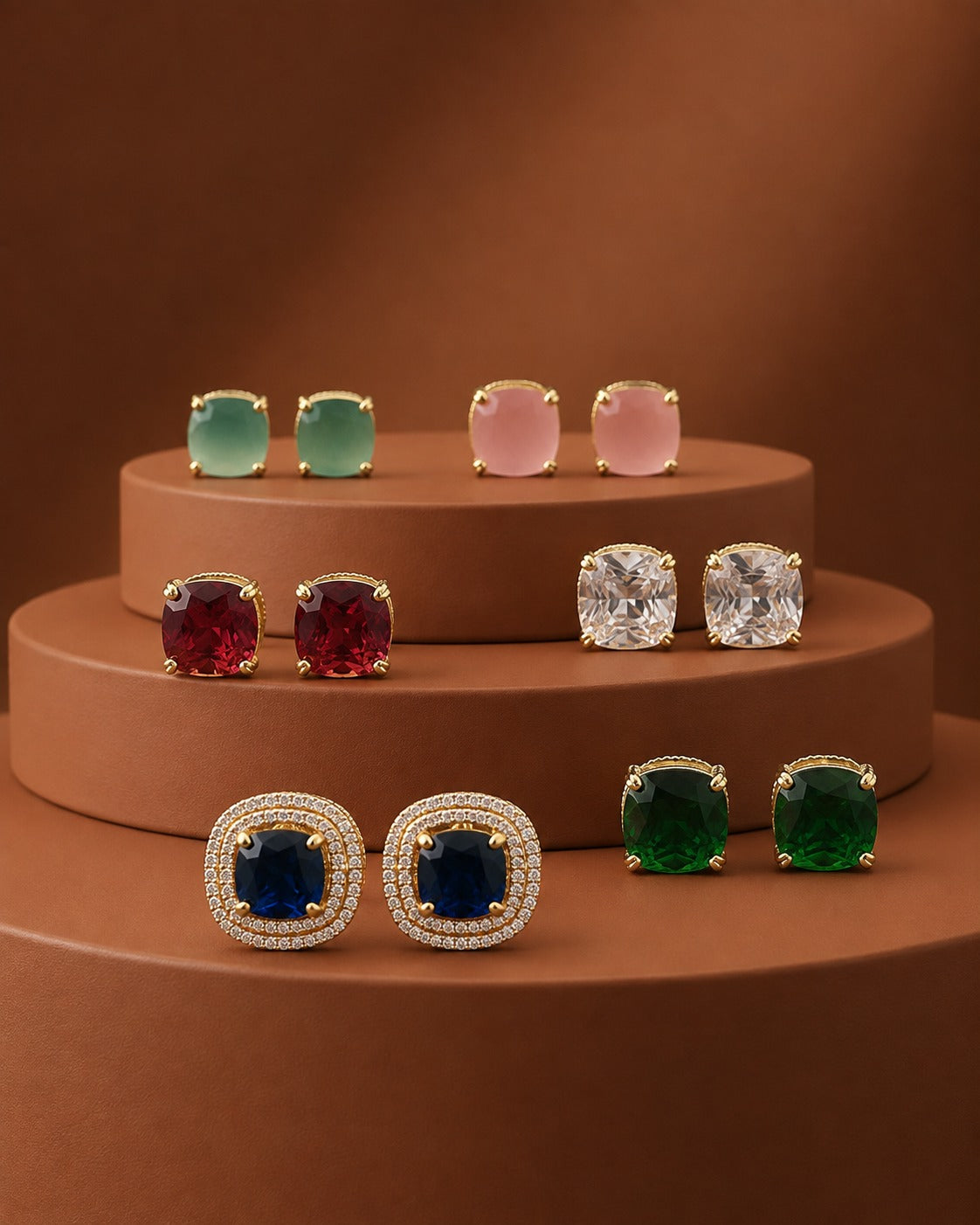 Six pairs of square stud earrings in gold settings, displayed on a tiered brown pedestal. The earrings feature various colored stones: mint green, light pink, deep red, clear, deep blue, and emerald green.