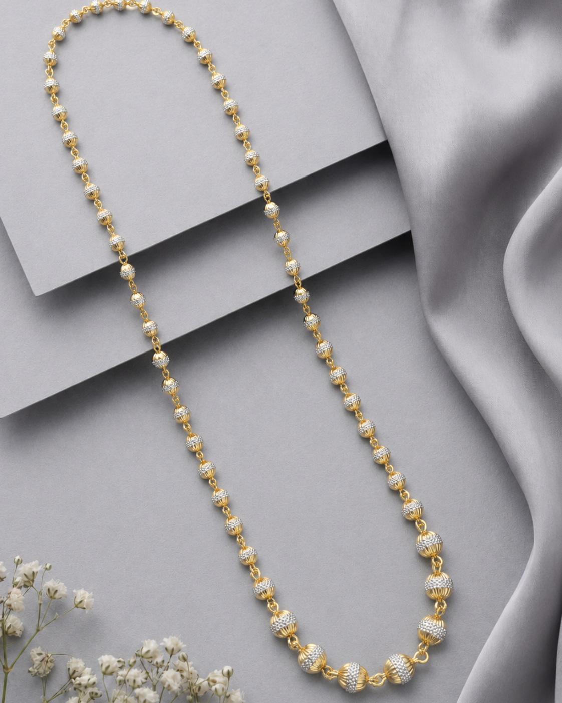A gold-toned beaded necklace with alternating smooth round beads and textured barrel-style beads, displayed on a light gray surface with draped satin fabric and small white flowers.