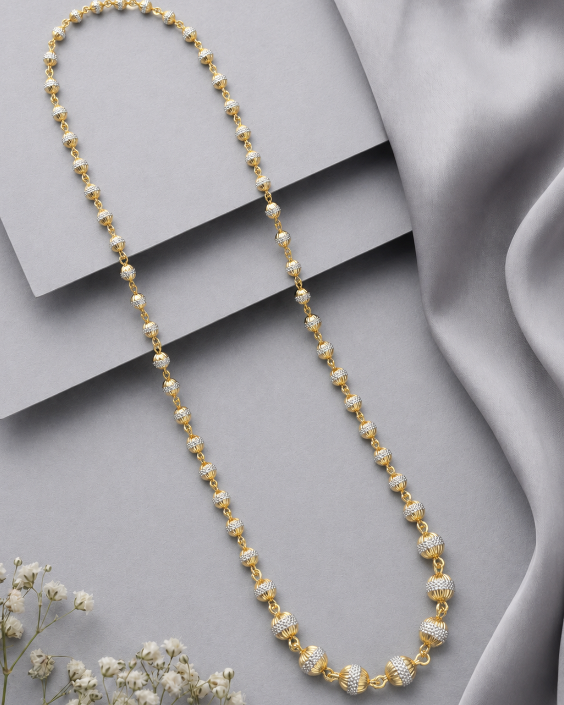 A gold-toned beaded necklace with alternating smooth round beads and textured barrel-style beads, displayed on a light gray surface with draped satin fabric and small white flowers.