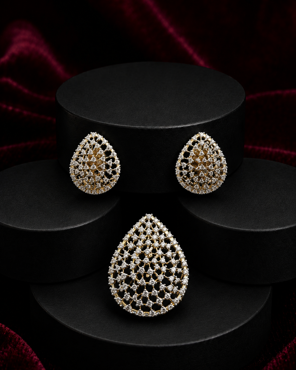 Teardrop-shaped jewelry set featuring a matching pair of earrings and a larger pendant or brooch, both adorned with intricate pave-set clear stones in a gold-tone setting, displayed on black cylindrical pedestals against a dark red velvet background.
