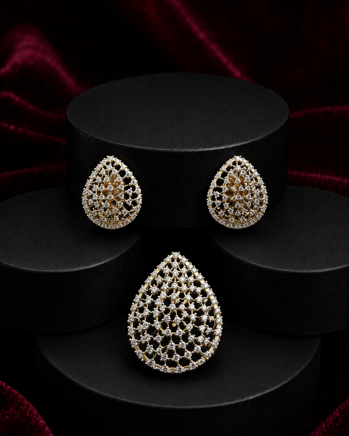 Teardrop-shaped jewelry set featuring a matching pair of earrings and a larger pendant or brooch, both adorned with intricate pave-set clear stones in a gold-tone setting, displayed on black cylindrical pedestals against a dark red velvet background.