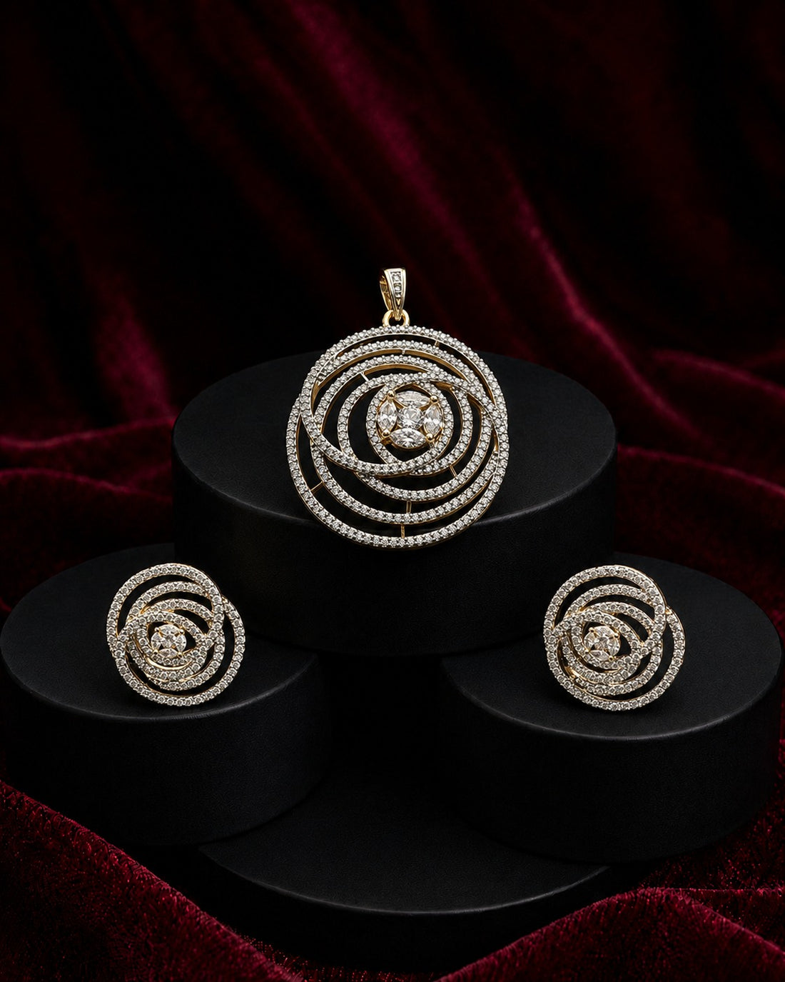 A jewelry set featuring a spiral pendant, matching spiral earrings, and a small matching charm, all set in a yellow-gold tone and adorned with clear stones, displayed on black cylindrical pedestals against a dark red velvet background.