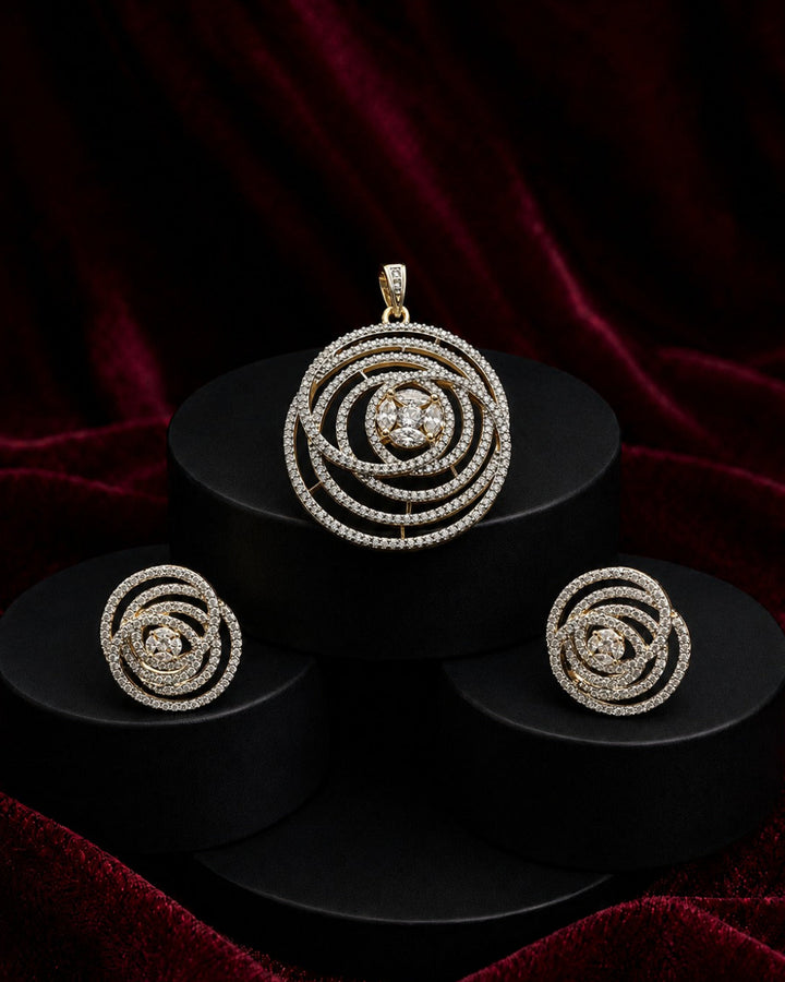 A jewelry set featuring a spiral pendant, matching spiral earrings, and a small matching charm, all set in a yellow-gold tone and adorned with clear stones, displayed on black cylindrical pedestals against a dark red velvet background.
