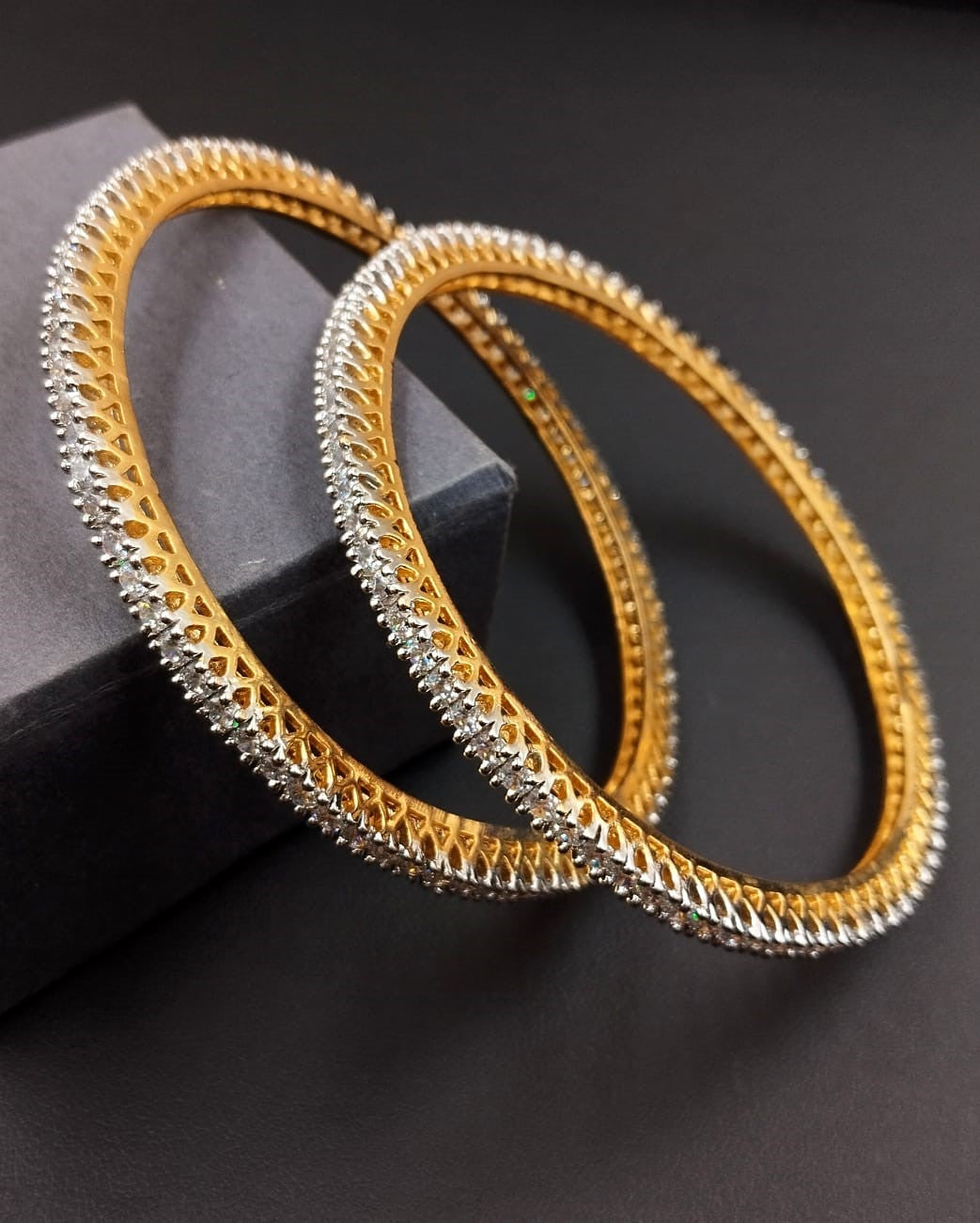 Two gold bangles with intricate designs on a dark surface