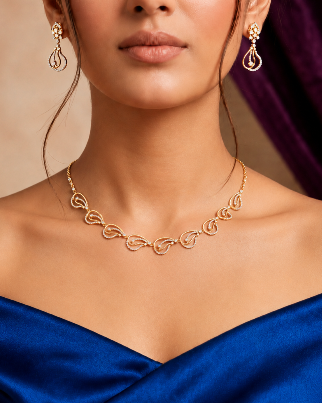 A close-up of a woman wearing a delicate gold necklace and matching drop earrings, styled with a royal blue satin dress against a warm beige background with a dark purple drape.