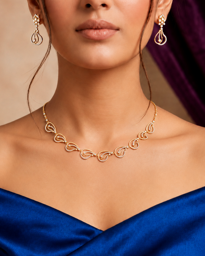 A close-up of a woman wearing a delicate gold necklace and matching drop earrings, styled with a royal blue satin dress against a warm beige background with a dark purple drape.