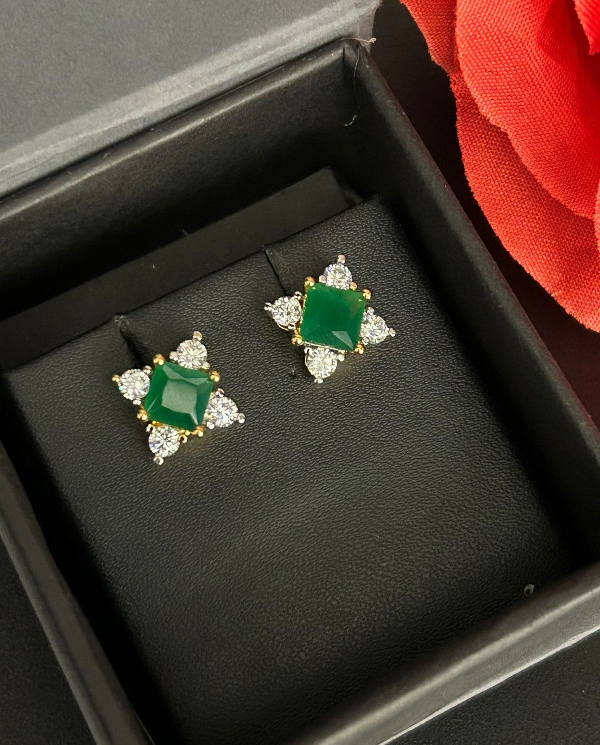 Pair of emerald and diamond earrings in a black jewelry box with a red flower in the background.