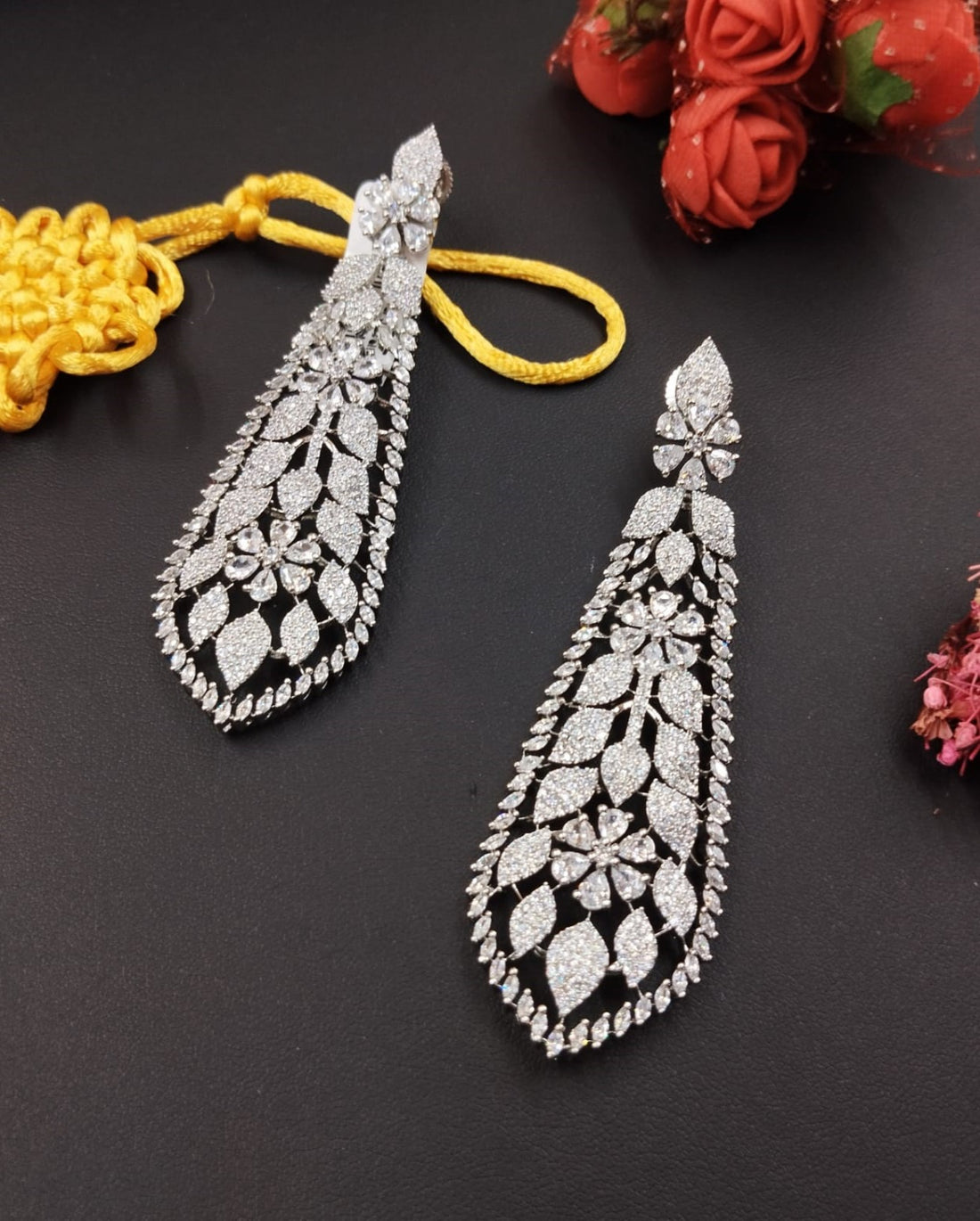 Two silver teardrop-shaped earrings on a dark surface with a yellow thread and red flowers.