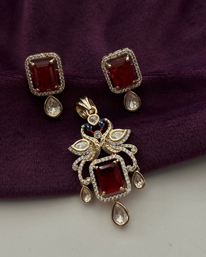 Gold-plated jewelry set featuring a matching pendant and stud earrings with rectangular deep red gemstones, surrounded by clear crystals and small blue accent stones, displayed on a textured dark purple fabric background.