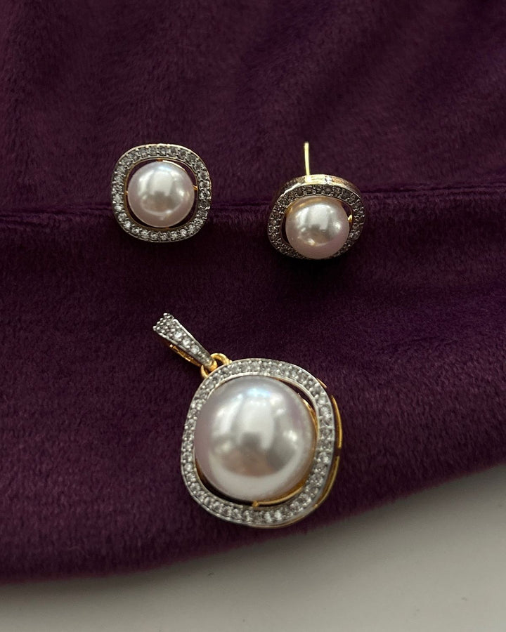 A jewelry set featuring a pendant and matching stud earrings with large white pearls surrounded by clear pavé stones in a gold-tone setting, displayed on a textured dark purple fabric background.