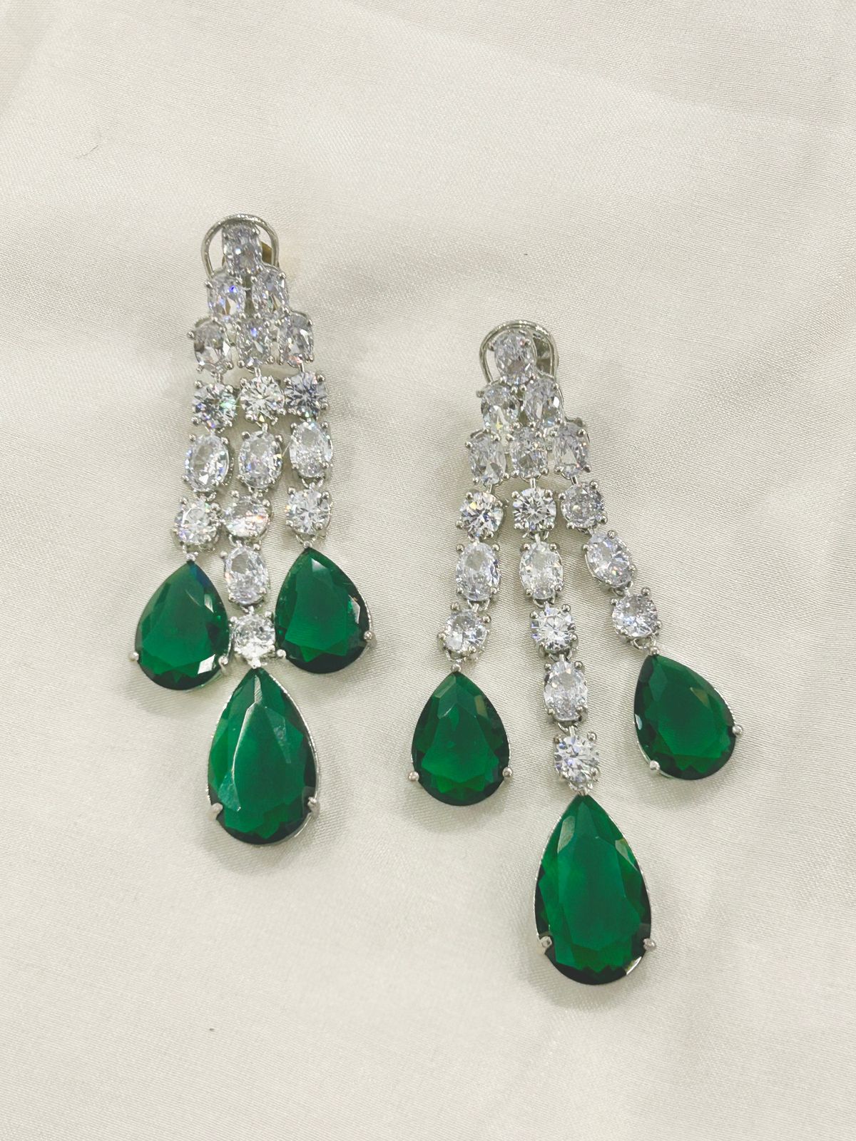 A pair of silver-tone chandelier earrings featuring three cascading rows of clear round-cut stones and three teardrop-shaped emerald green gemstones at the bottom, displayed on a light beige fabric background.