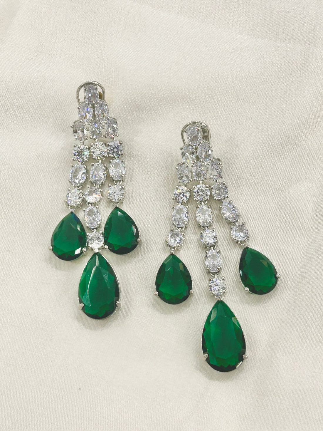 A pair of silver-tone chandelier earrings featuring three cascading rows of clear round-cut stones and three teardrop-shaped emerald green gemstones at the bottom, displayed on a light beige fabric background.