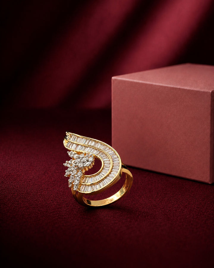 A gold ring with a curved, wing-like design paved with clear stones, displayed on a dark red velvet surface next to a dusty rose jewelry box.