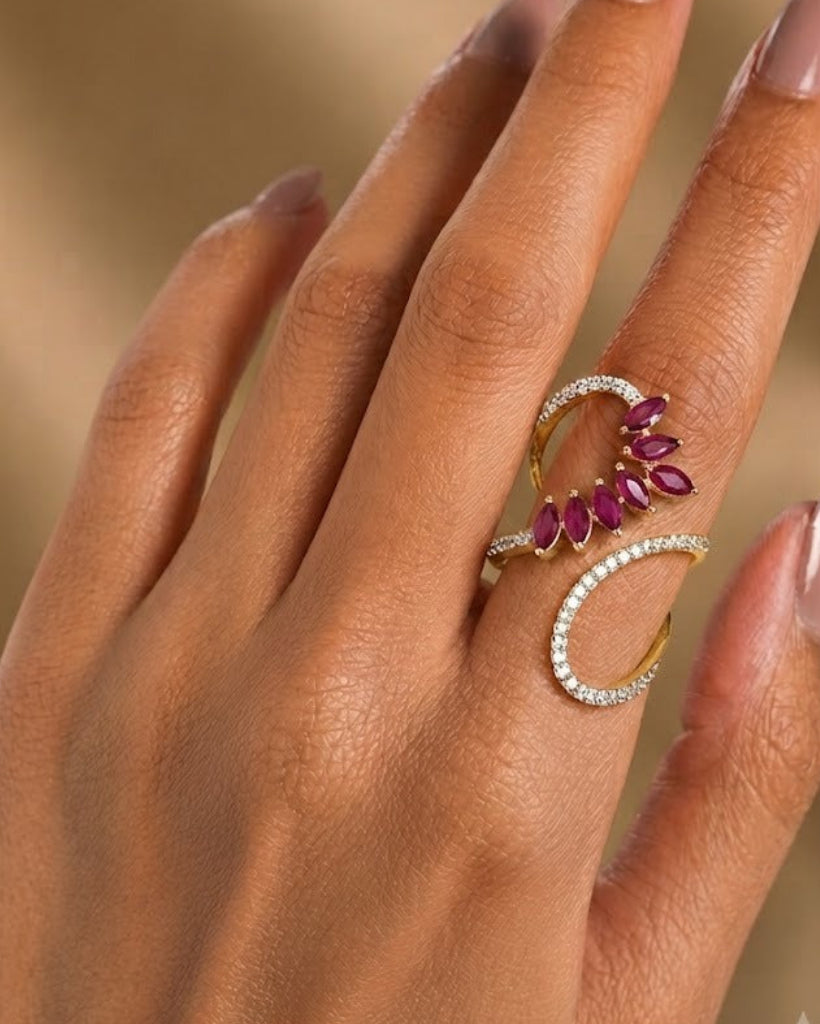 A close-up of a hand wearing a gold ring with a double crescent design, one half encrusted with small clear stones and the other featuring a row of deep pink gemstones.