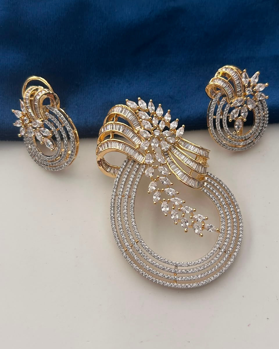 A gold and silver-toned jewelry set featuring a large teardrop-shaped brooch with a matching pair of earrings, both adorned with clear pavé stones, displayed on a white surface with a dark blue fabric background.