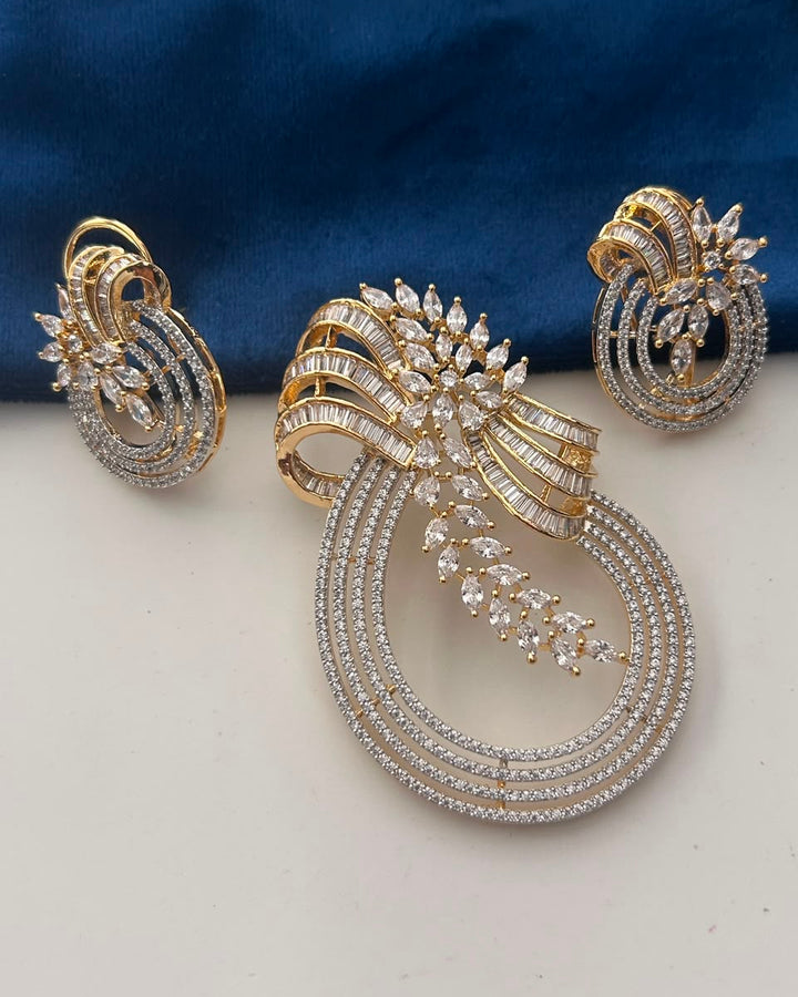 A gold and silver-toned jewelry set featuring a large teardrop-shaped brooch with a matching pair of earrings, both adorned with clear pavé stones, displayed on a white surface with a dark blue fabric background.
