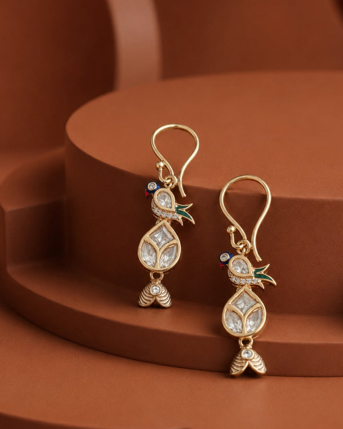 A pair of gold-toned dangle earrings with clear stone leaf motifs and small green and blue enamel accents, displayed on a terracotta-colored geometric background.