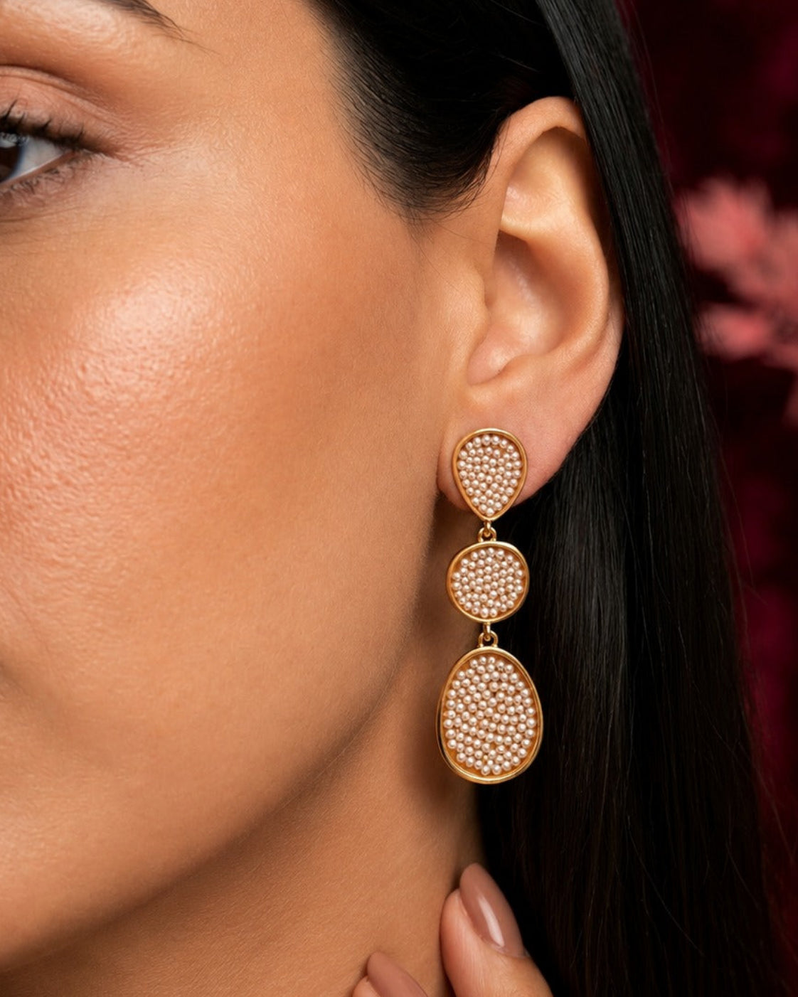 A close-up of a woman wearing a gold-toned dangle earring with three stacked oval clusters of small clear stones against a dark red background.