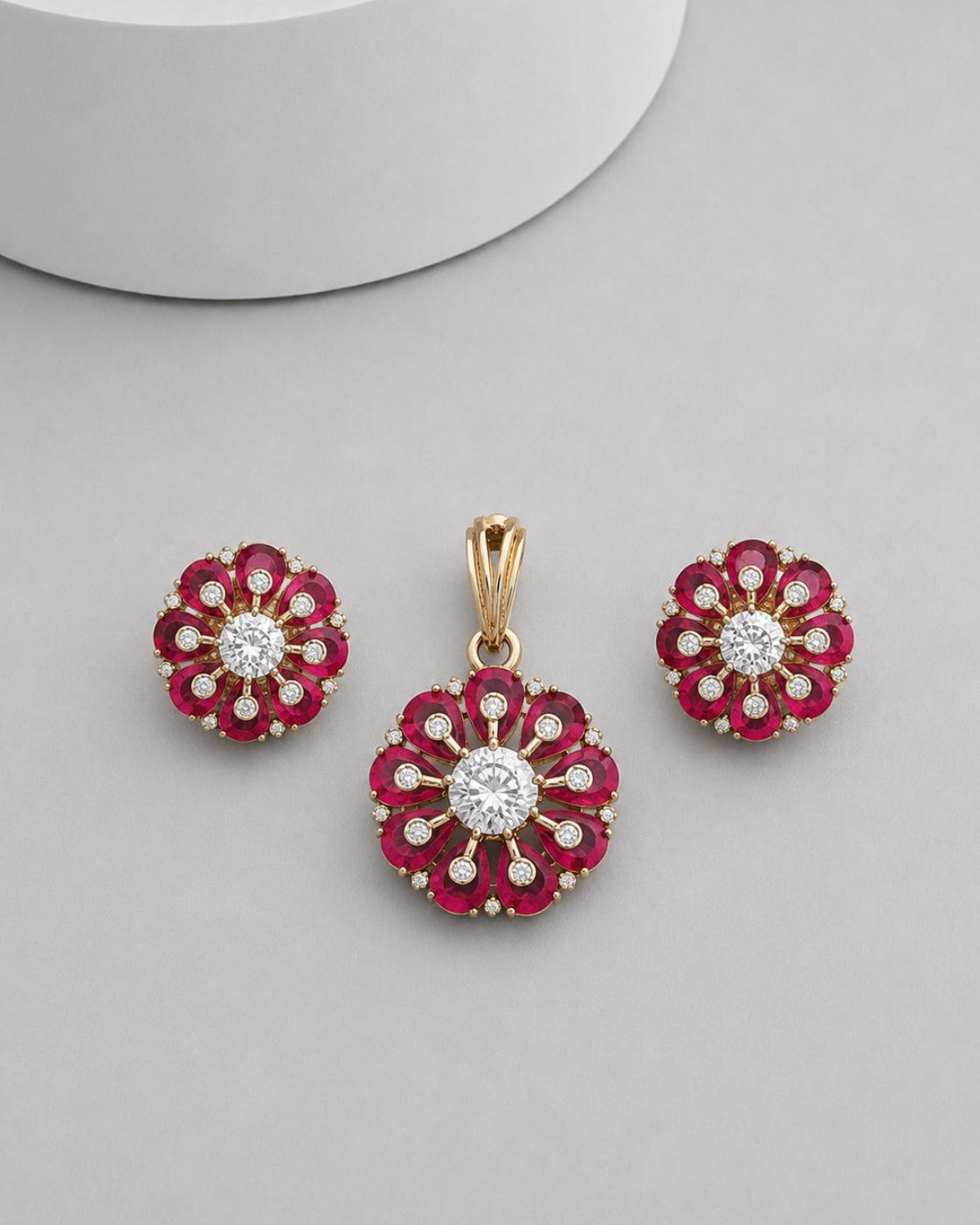A gold flower-shaped pendant and matching stud earrings set with deep red gemstones and clear center stones, displayed on a light gray surface.