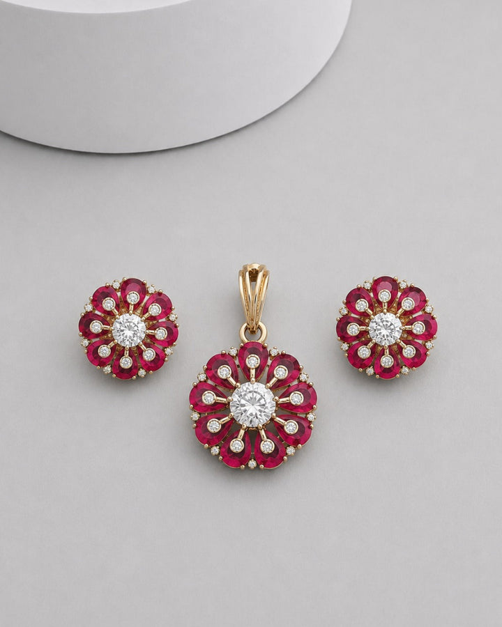 A gold flower-shaped pendant and matching stud earrings set with deep red gemstones and clear center stones, displayed on a light gray surface.