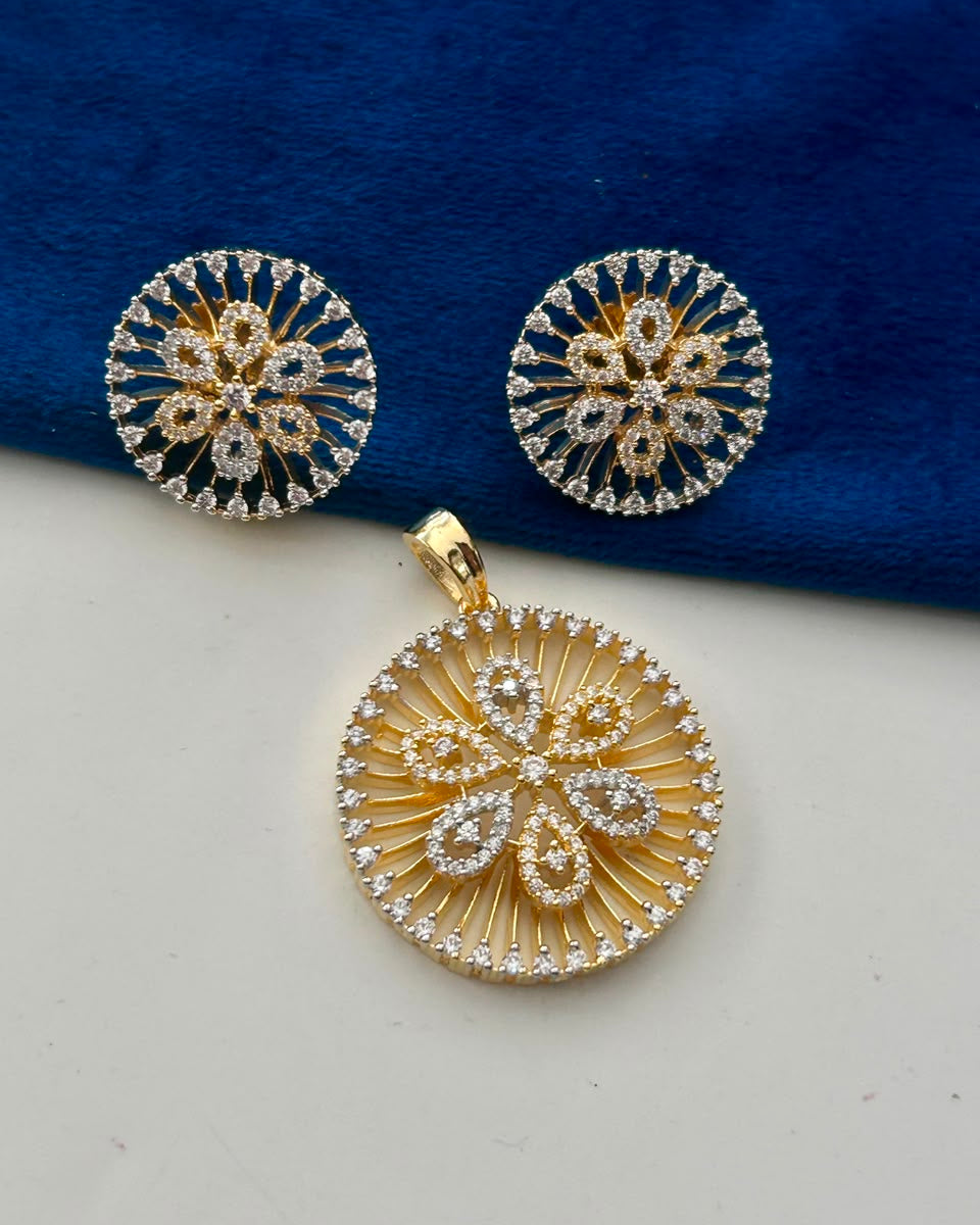 Gold-toned jewelry set featuring a round pendant and matching stud earrings with intricate floral and sunburst designs set with clear stones, displayed on a split white and dark blue background.